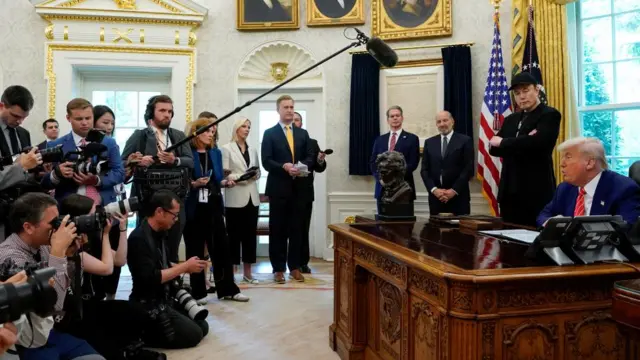 Elon Musk and Donald Trump speak in the Oval Office in front of a group of reporters, holding microphones and cameras.