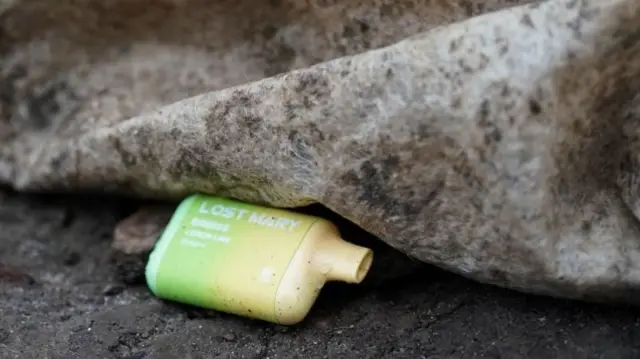 A vape lying on a dirty floor next to a dirty bit of carpet