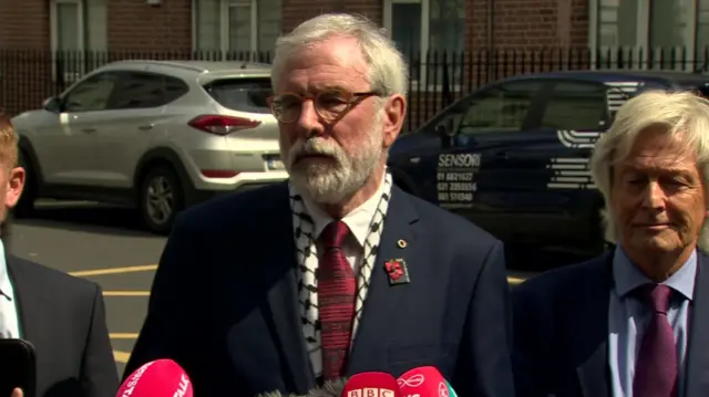 A grey haired man with a navy suit and red tie stands in front of 5 microphones, he wears brown glasses.