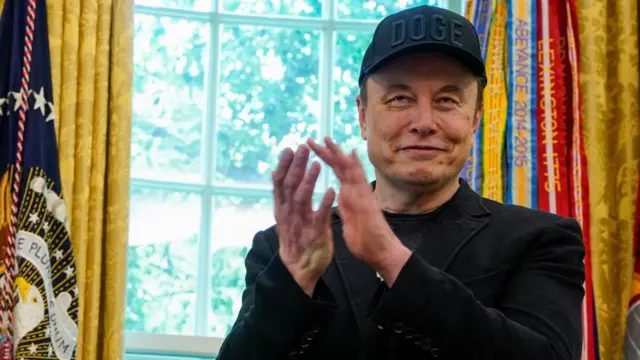 Elon wears a black cap reading: 'Doge'. He also wears a black top and blazer, and claps his hands in the Oval Office.
