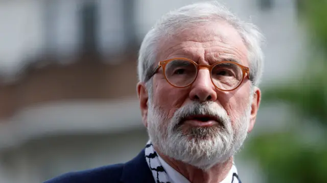 A grey haired man with brown glasses, has grey facial hair and navy suit. He also wears a white shirt.