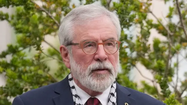 Gerry has grey hair, beard and orange glasses. He is wearing a navy suit and white shirt and red tie. Trees are behind him.
