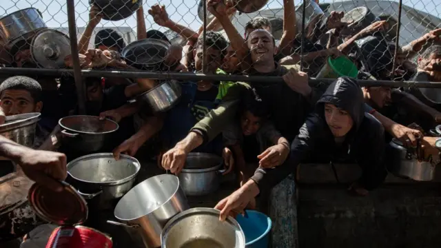 People hold out their pots through a gap in the fence, hoping to receive food from a person holding a large tin. The crowd appears to be squashed against the fence as people try and get a place