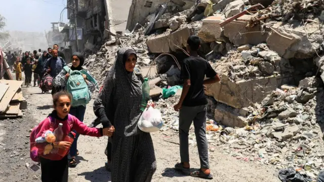 Palestinians evacuate in the aftermath of an Israeli strike in Gaza city. Woman, some holding the hands of children, walk alongside rubble and smoke.