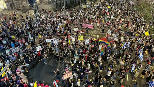 Demonstrators take part in a protest against Israeli Prime Minister Benjamin Netanyahu's government. a crowd of at least 1,000 people is gathered, they are holding yellow signs and fill a street. the photo is an aerial photo showing the crowd