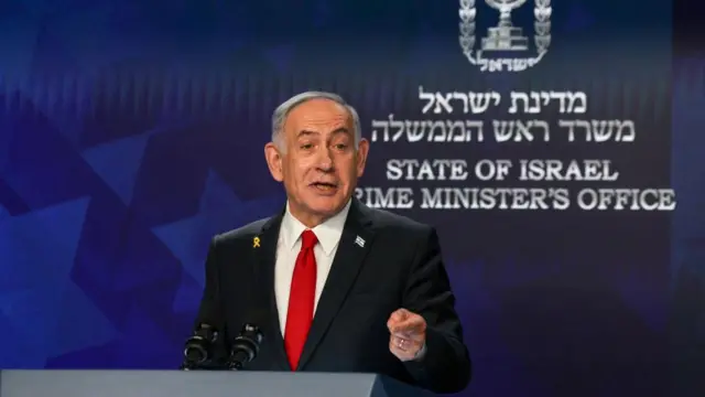 Israeli Prime Minister Benjamin Netanyahu speaks during a press conference wearing a blazer and red tie, in front of a backdrop reading State of Israel Prime Minister's Office.
