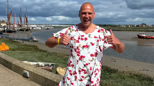 A man smiling, dressed in a white dress with red flowers, with his thumbs up