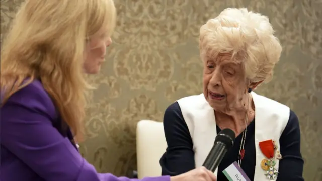 Elderly woman with platinum blonde hair sitting down on a chair wearing a black and white cardigan and black shirt as she gives an interview to a blonde woman in a purple suit