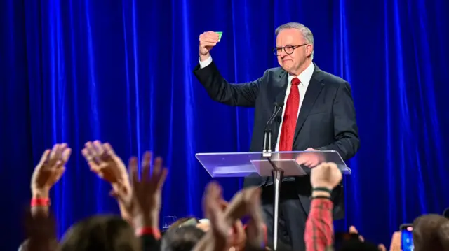 Prime Minister Anthony Albanese wears a red tie and suit and holds up a green card to a cheering crowd.