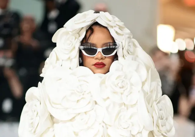 Rihanna at the Met Gala, wears a white dress of roses that covers her head, red lipstick and white sunglasses with fake eyelashes stuck on them.