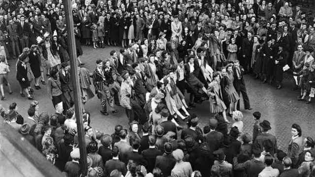 A large crowd of men and women observe as others dance in the middle of the street