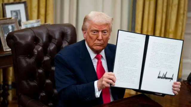 Trump holds an executive order, signed by him, in the Oval Office wearing a suit and red tie.