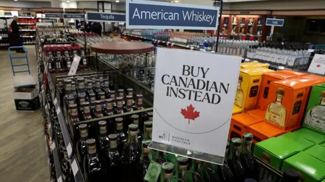 A sign that reads ''Buy Canadian Instead'' is displayed on top of bottles, hanging above another sign that reads "American Whiskey" in a store.