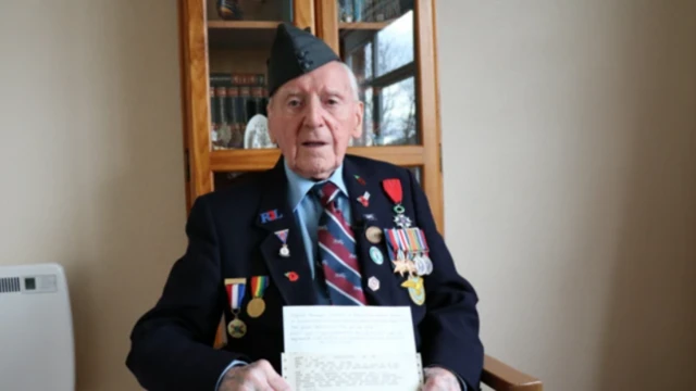 An elderly veteran sits down in a wooden chair wearing a dark jacket covered in multiple military medals on both sides of his chest. He's donning a dark grey carp. Behind him is a wooden bookcase with glass windows