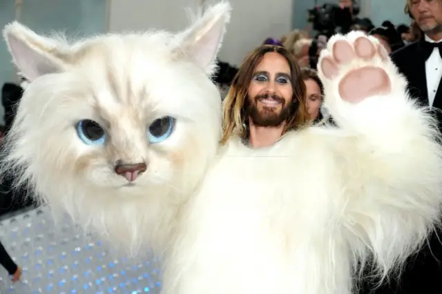 Jared Leto in a huge and very realistic cat outfit, waving his paw and holding the head part of the cat outfit at the 2023 Met Gala.