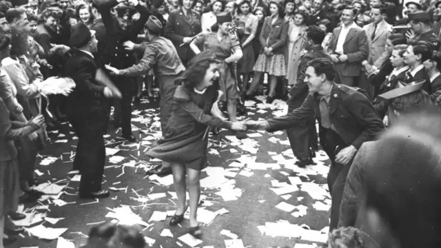 A young woman with long brown hair in a dark dress and wearing a dark military side cap laughs as she dances in the street with a young brown-haired man in uniform, who's also smiling. Around them is a large crowd