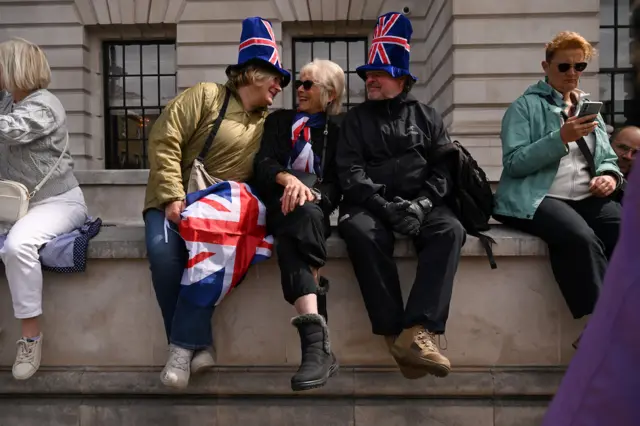 Three people perch holding a British flag and wearing large hats with the British flag on