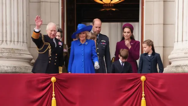 King Charles and Queen Camilla, Princess Anne, the Prince and Princess of Wales and their children stand on the balcony at Buckingham Palace - the King waves