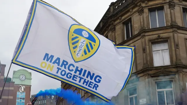 A Leeds flag which reads Marching On Together waved in the air next to a smoking blue flare.