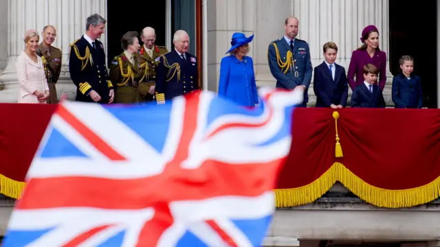 Wide shot of all the royals on balcony with UK flag waving in the foreground