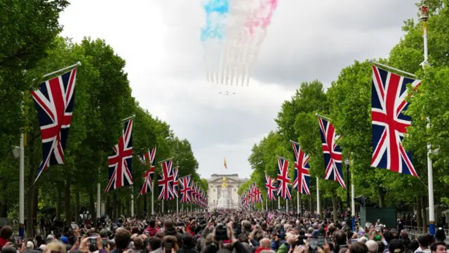 A large crowd fills the Mall, which is lined with Union Jack flags, as the Red Arrows past overhead, followed by red, white and blue smoke