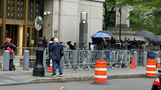 Media members are enclosed in metal barricades with cameras and tripods. Several umbrellas are up.