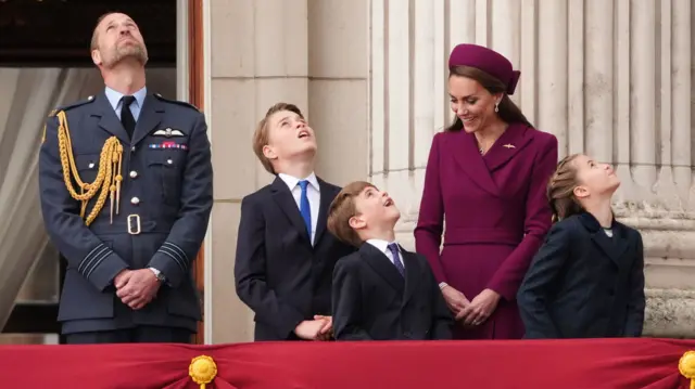 The Prince and Princess of Wales and their children look up into the air as the flypast goes past