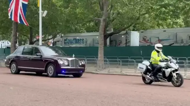 Queen in car with motorcycle in front
