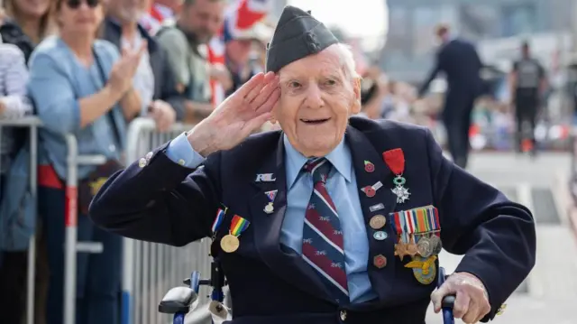 Veteran Bernard Morgan, wearing a suit with several medals pinned to it, salutes, with the VE Day crowd behind him