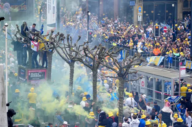 Fans line either side of a road through Leeds city centre. Several yellow flares have been lit.