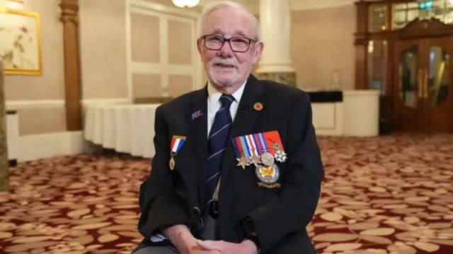 Alan Kennett smiles as he sits wearing a suit with medals pinned to his jacket