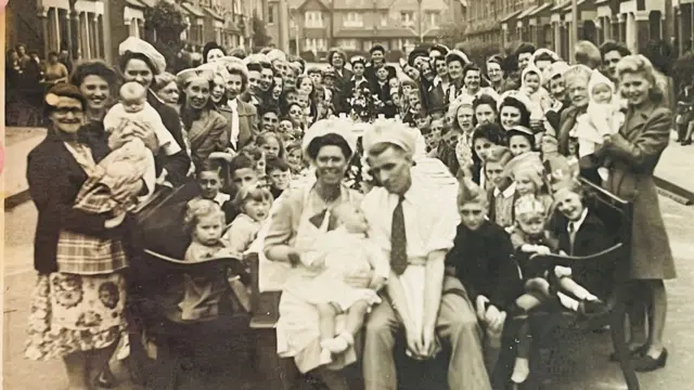 A black and white photo of a street party - children are seated at a long table while adults stand around the outside