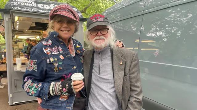 Jenny Sirell has her arm around Richard Singleton and they smile at the camera. A coffee cart can be seen behind them