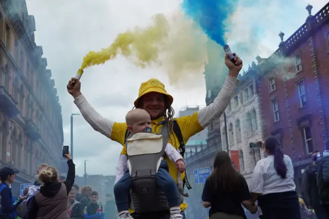 A man holds a yellow and blue flare aloft. He is wearing a baby carrier, carriyng a small child