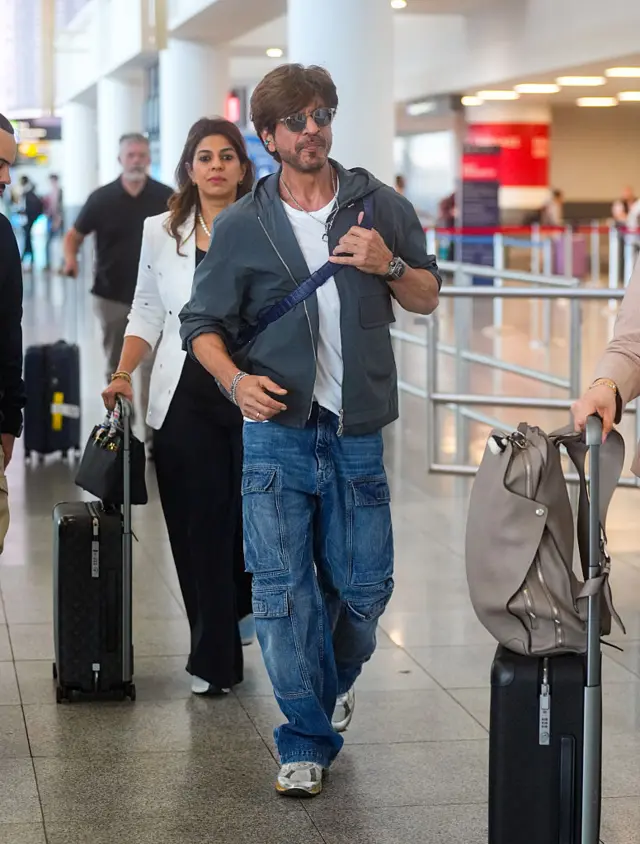 Shah Rukh Khan walks at an airport. He is wearing a teal jacket over a white shirt and blue jeans. He is wearing sunglasses and a blue leather bag is strapped across his torso.