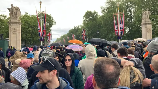 Crowds walking through Buckingham Palace gates with umbrellas and hoods