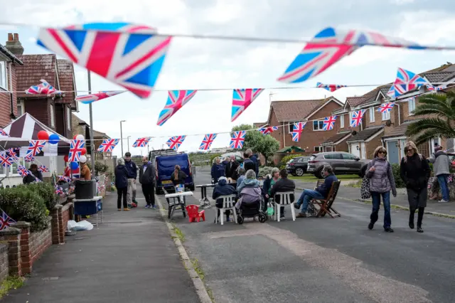 People sat on plastic chairs in the road covered with Union Jack flags