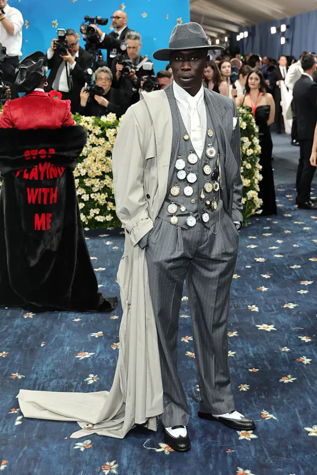 Khaby Lame in a grey stripped suit and trousers over a white shirt. His grey petticoat is decorated with several pocketclocks.