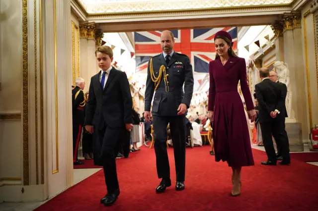 Catherine, Prince William, and Prince George walk on red carpet with UK flag hanging from the ceiling behind them