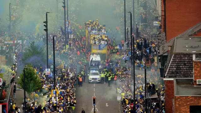 The buses make their way down a packed street with a police escort.