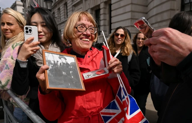 Wearing a red jacket and spactacles, a woman holds a picture of her veteran father as people gather to watch an armed forces and veterans' parade