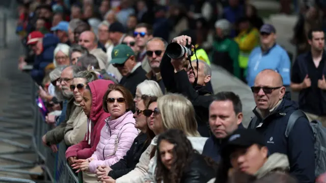 Lines of people watch on behind metal barriers