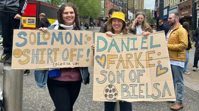 Two women holding cardboard signs which read 'Solomon shirt off' and 'Daniel Farke: son of Bielsa'.