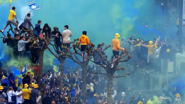 People on trees and the street below against a backdrop of smoke from flares