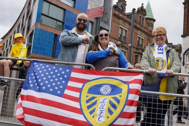 A man and a woman stood next to a flag of America with the Leeds United logo printed on. Matt is wearing sunglasses and a Leeds United shirt, while Valerie is wearing a Leeds United scarf.
