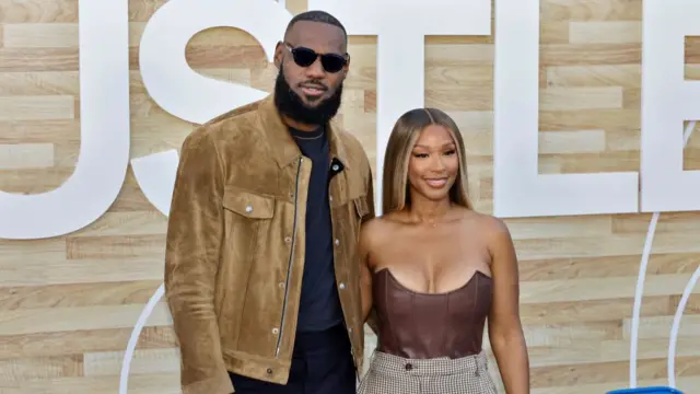 LeBron James and his wife Savannah on a red carpet. He is wearing a brown jacket over a black shirt and trousers and he is wearing black sunglasses. Savannah James is wearing a strapless leather shirt and chequered trousers.