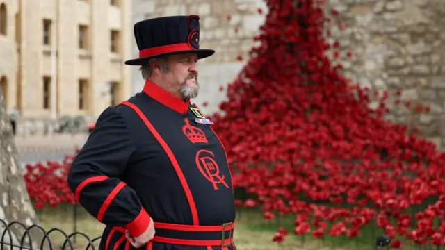 A Yeoman Warder in his uniform - black and red with a hat - stands next to a display ceramic poppies