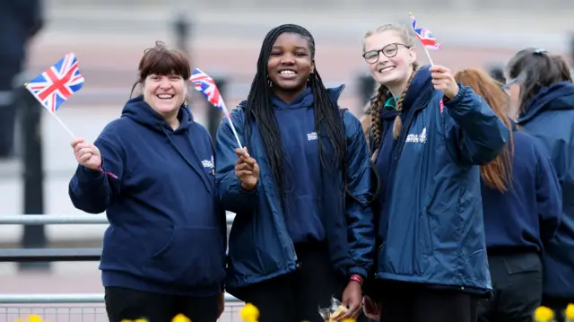 Three women wearing navy hoodies smile at the camera while waving UK flags