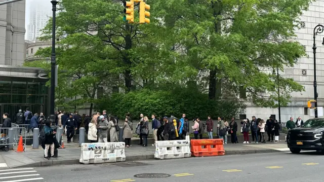 People line up outside a court building in New York City. Large green trees are behind them and there are some orange and white barricades in front.