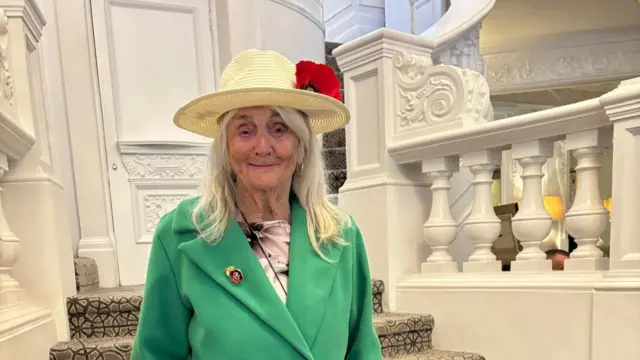 A headshot of Joan Poppy Taylor wearing a green coat and a poppy in her hat